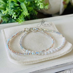 May include: Two delicate bracelets featuring iridescent crystal beads and smooth white round beads. The jewelry includes silver-toned metal spacers and lobster claw clasps. The text J Grace Jewelry is visible above the bracelets on a soft fabric.