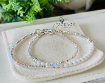 May include: Two delicate bracelets featuring iridescent crystal beads and smooth white round beads. The jewelry includes silver-toned metal spacers and lobster claw clasps. The text J Grace Jewelry is visible above the bracelets on a soft fabric.