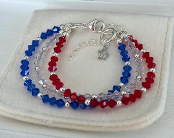 Puede incluir: Tres pulseras de cuentas rojas, blancas y azules, sobre una tela blanca. Las pulseras tienen cierres plateados y un pequeño dije de estrella. Las cuentas son facetadas y están dispuestas en filas.