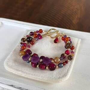 May include: A multi-strand bracelet featuring a toggle clasp. The bracelet is composed of various gemstone beads in shades of red, purple, orange, and brown. Gold-colored beads and accents are interspersed throughout the design. The bracelet rests on a white surface.