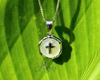 May include: A silver-toned necklace featuring a circular pendant with a white, iridescent inlay and a centered silver cross. The pendant hangs from a delicate box chain. The text J GRACE JEWELRY is printed in dark blue below the pendant.