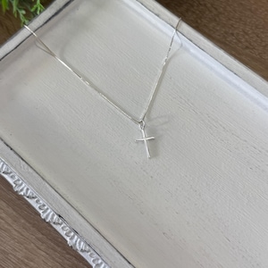 May include: A delicate silver-toned necklace featuring a thin chain and a small, simple cross pendant. The metal has a polished, reflective finish, and the cross is suspended from the center of the chain, creating a clean and understated appearance.