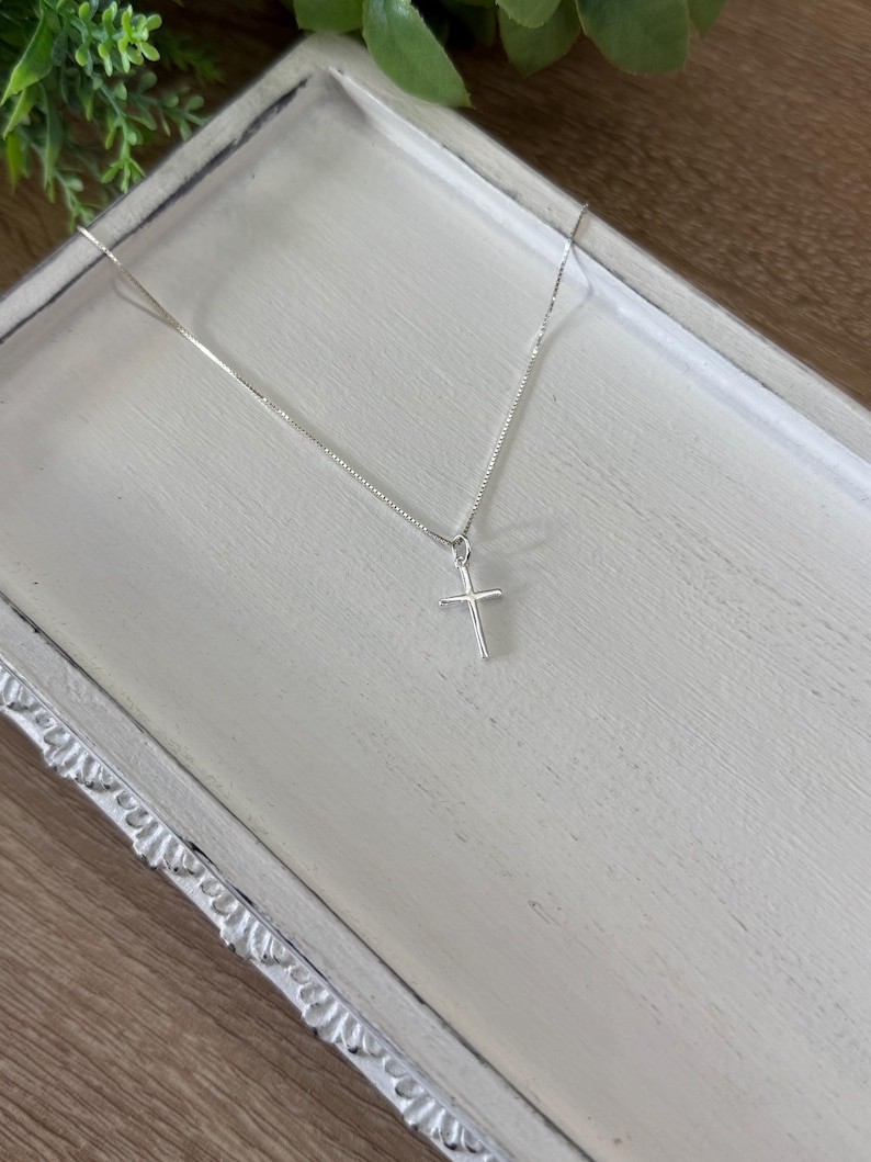 May include: A delicate silver-toned necklace featuring a thin chain and a small, simple cross pendant. The metal has a polished, reflective finish, and the cross is suspended from the center of the chain, creating a clean and understated appearance.