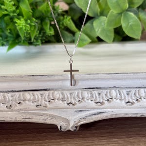 May include: A silver-toned metal cross pendant suspended from a delicate, thin box chain. The cross features a simple, smooth design with a polished finish, hanging vertically from a small jump ring attached to the slender metallic necklace chain.