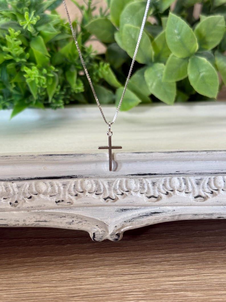 May include: A silver-toned metal cross pendant suspended from a delicate, thin box chain. The cross features a simple, smooth design with a polished finish, hanging vertically from a small jump ring attached to the slender metallic necklace chain.