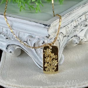 May include: A gold-colored necklace with a rectangular pendant. The pendant features a floral design with intricate details. The necklace chain is also gold and has a delicate, rectangular link pattern. The background is a blurred white surface.