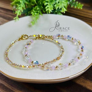 May include: Two delicate bracelets featuring clear iridescent crystal beads, small light pink round beads, and gold-toned metal accents. The bracelets have gold-toned lobster claw clasps. The text J Grace Jewelry is visible in the upper right.
