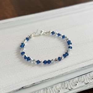 May include: A delicate bracelet featuring alternating blue and clear crystal beads strung on a silver chain. The bracelet is displayed on a white, distressed wooden surface, showcasing its elegant design and craftsmanship.