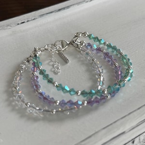 May include: Three delicate bracelets with iridescent crystal beads in shades of clear, teal, and lavender. The bracelets are strung on a silver chain with a lobster clasp. The jewelry is displayed on a white surface.