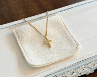 May include: A gold-colored cross pendant necklace displayed on a white, rectangular, soft cloth. The necklace chain is delicate, and the cross is a simple, classic design. The background is a distressed white tray with a decorative edge.
