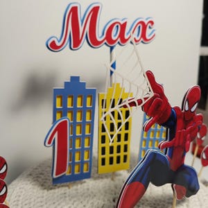 Puede incluir: Un conjunto de adornos para tartas con tem&aacute;tica de Spider-Man. El conjunto incluye una figura de Spider-Man, el n&uacute;mero 1 y edificios de la ciudad. El nombre "Max" se muestra en rojo y azul. La combinaci&oacute;n de colores es rojo, azul y amarillo.