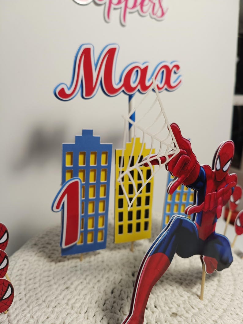 Puede incluir: Un conjunto de adornos para tartas con tem&aacute;tica de Spider-Man. El conjunto incluye una figura de Spider-Man, el n&uacute;mero 1 y edificios de la ciudad. El nombre "Max" se muestra en rojo y azul. La combinaci&oacute;n de colores es rojo, azul y amarillo.