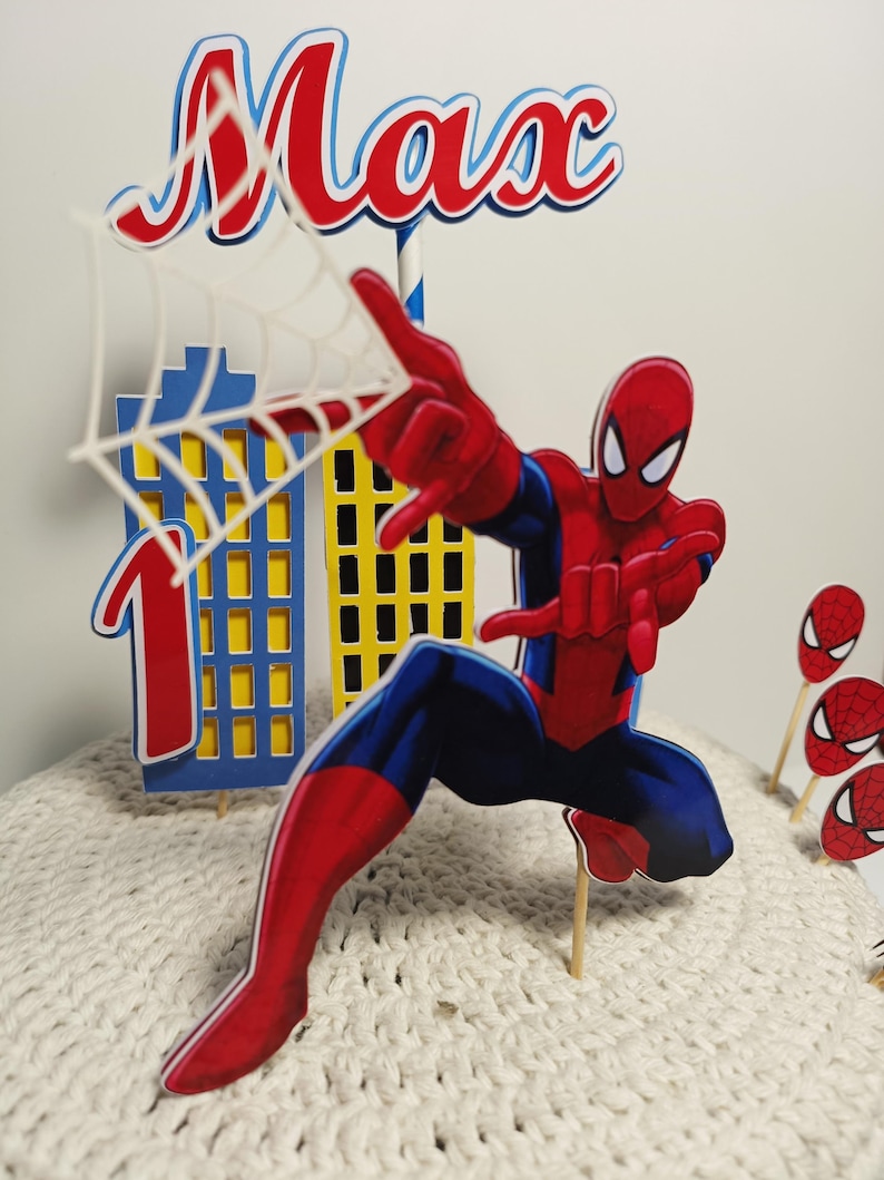Puede incluir: Un adorno para tarta con tem&aacute;tica de Spider-Man en rojo, azul y blanco, con el nombre "Max" en letras rojas. El dise&ntilde;o incluye un paisaje urbano, el n&uacute;mero "1" y varios recortes de caras de Spider-Man. La figura principal es Spider-Man en una pose din&aacute;mica.