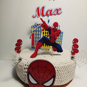 Puede incluir: Un juego de adornos para tartas con tem&aacute;tica de Spider-Man. El juego incluye una figura de Spider-Man, el nombre "Max", el n&uacute;mero "1" y varias decoraciones de telara&ntilde;as y cabezas de ara&ntilde;a. La tarta est&aacute; cubierta con una tela de ganchillo blanca.