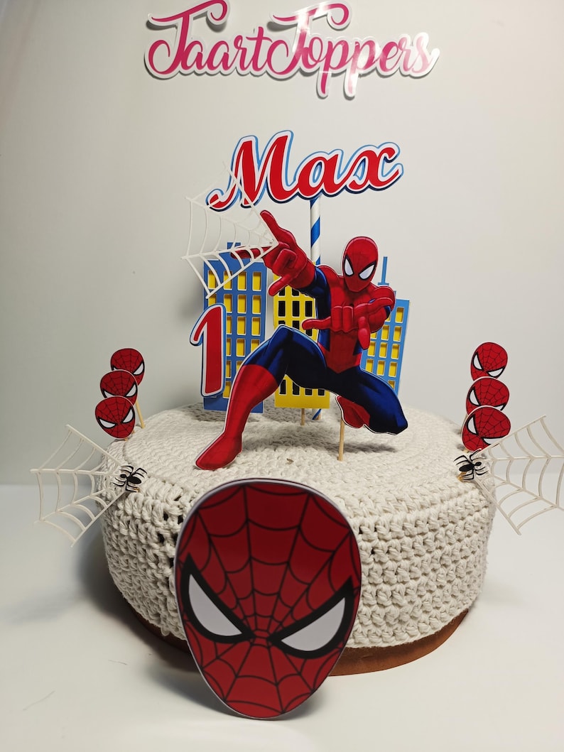 Puede incluir: Un juego de adornos para tartas con tem&aacute;tica de Spider-Man. El juego incluye una figura de Spider-Man, el nombre "Max", el n&uacute;mero "1" y varias decoraciones de telara&ntilde;as y cabezas de ara&ntilde;a. La tarta est&aacute; cubierta con una tela de ganchillo blanca.
