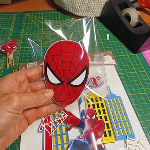 Puede incluir: Un juego de decoraci&oacute;n para fiestas con tem&aacute;tica de Spider-Man. El juego incluye una m&aacute;scara de Spider-Man roja y negra, una figura de Spider-Man y recortes de edificios. La m&aacute;scara est&aacute; en una bolsa de pl&aacute;stico transparente.
