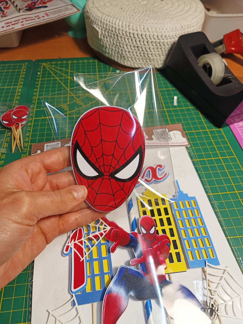 Puede incluir: Un juego de decoraci&oacute;n para fiestas con tem&aacute;tica de Spider-Man. El juego incluye una m&aacute;scara de Spider-Man roja y negra, una figura de Spider-Man y recortes de edificios. La m&aacute;scara est&aacute; en una bolsa de pl&aacute;stico transparente.