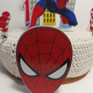Puede incluir: Adorno para tarta con m&aacute;scara de Spider-Man roja y negra con ojos blancos y dise&ntilde;o de telara&ntilde;a. Otras decoraciones incluyen un n&uacute;mero uno, una figura de Spider-Man y recortes de horizonte. La tarta est&aacute; sobre una superficie blanca.