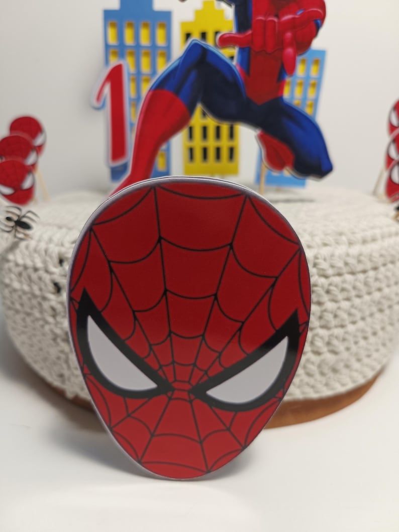 Puede incluir: Adorno para tarta con m&aacute;scara de Spider-Man roja y negra con ojos blancos y dise&ntilde;o de telara&ntilde;a. Otras decoraciones incluyen un n&uacute;mero uno, una figura de Spider-Man y recortes de horizonte. La tarta est&aacute; sobre una superficie blanca.