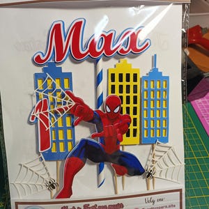 Puede incluir: Adorno de pastel colorido con el nombre "Max" en rojo y azul, y una figura de Spider-Man. El dise&ntilde;o incluye edificios de la ciudad, telara&ntilde;as y ara&ntilde;as. El texto "Taarttoppers" es visible.