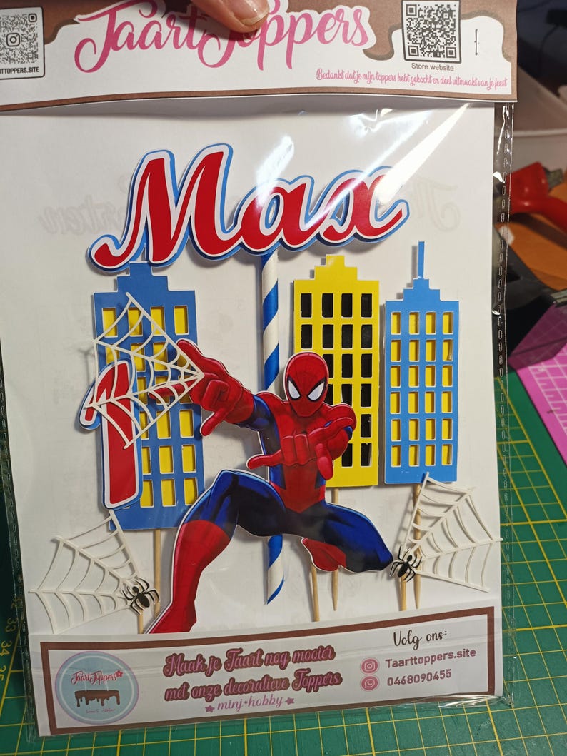 Puede incluir: Adorno de pastel colorido con el nombre "Max" en rojo y azul, y una figura de Spider-Man. El dise&ntilde;o incluye edificios de la ciudad, telara&ntilde;as y ara&ntilde;as. El texto "Taarttoppers" es visible.