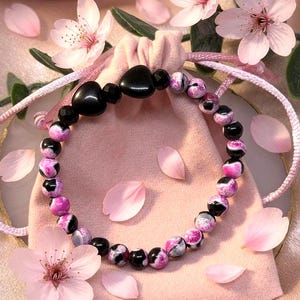 Può includere: Un braccialetto di perline con perline nere a forma di cuore e perline rotonde con motivi rosa e neri. Il braccialetto è su un sacchetto rosa con coulisse, circondato da petali e fiori rosa.