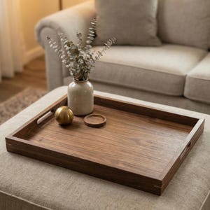 May include: A rectangular wooden serving tray with integrated handles, showcasing a rich, dark wood grain. A small, round coaster and a gold sphere sit on the tray, alongside a small vase with dried foliage. The tray rests on a neutral-toned ottoman.
