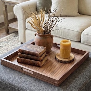 May include: A wooden serving tray with a warm brown finish, holding a stack of antique books, a terracotta vase with dried plants, and a yellow candle on a wooden coaster. The tray is rectangular with cut-out handles.