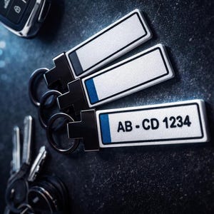 Könnte beinhalten: Drei rechteckige Schlüsselanhänger mit weißer Oberfläche und schwarzem Rand, einer mit der Aufschrift "AB-CD 1234" in schwarzer Schrift. Jeder Schlüsselanhänger hat einen blauen Streifen und einen silbernen Schlüsselring. Ein Autoschlüssel und andere Schlüssel sind ebenfalls sichtbar.