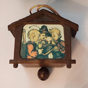 May include: A vintage wooden cuckoo clock ornament. The clock features a rectangular image of three children studying a chalkboard with the equation 1+1=, set within a brown wooden frame. A gold-colored cord is attached for hanging.