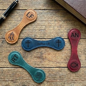 May include: An assortment of leather book markers in various colors, including tan, blue, teal, and red. Each marker features unique designs such as initials, a cross, a dove, a fish, and the text "Joshua 1:9". A pen and a book are also visible.