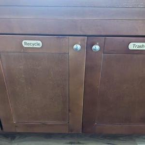 May include: Two wooden cabinet doors with silver knobs. The left door has a label that says "Recycle" and the right door has a label that says "Trash".