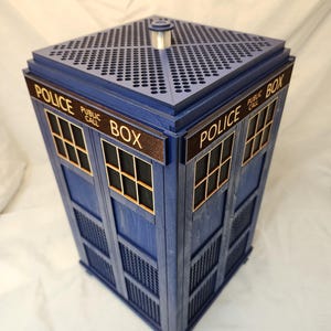 Peut inclure: Une boîte en bois bleue avec l'inscription "POLICE BOX" sur les côtés. Elle présente des fenêtres, un dessus perforé et un cylindre argenté. Le design rappelle les cabines téléphoniques britanniques.