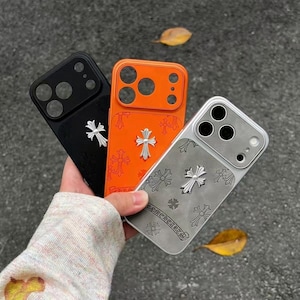 Könnte beinhalten: Drei Smartphone-Hüllen in Schwarz, Orange und Silber, die jeweils ein markantes silberfarbenes Kreuz-Emblem in der Mitte und kleinere Kreuzmuster auf der Rückseite aufweisen. Die Hüllen haben Aussparungen für Kameraobjektive und Blitz.