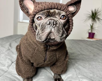 May include: A brown French Bulldog wearing a matching brown hooded sweater with ear-like extensions. The dog is sitting on a gray bedspread, looking directly at the camera. The sweater has a zipper closure.