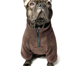 May include: A brown French Bulldog wearing a brown fleece dog sweater with a black zipper. The dog is looking up with a focused expression. The sweater has long sleeves and covers the dog's torso.