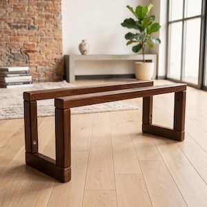 May include: A low-profile bench constructed from dark brown wood, featuring a rectangular frame design with four vertical support legs connected by horizontal beams, showcasing a smooth finish and visible joinery on the side of the frame.