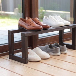 May include: A dark brown wooden shoe rack with two tiers. The top tier holds a pair of brown leather loafers and a pair of white sneakers. The bottom tier holds a pair of beige slip-on shoes and a pair of grey slip-on shoes with white soles.