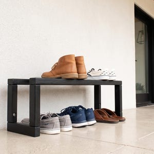 May include: A black wooden shoe rack with a rectangular frame and four legs. The top shelf holds a pair of tan leather boots and a pair of white slip-on sneakers. The bottom shelf holds a pair of gray sneakers, blue sneakers, and brown sandals.