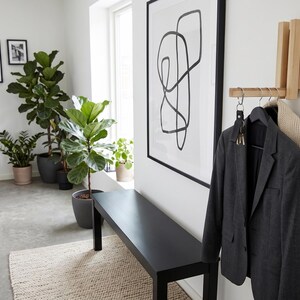 Könnte beinhalten: Eine wandmontierte Garderobe aus hellem Holz mit drei Metallhaken. Ein dunkelgrauer Blazer und ein cremefarbener Strickschal hängen an den Haken, wobei ein Schlüsselbund an einem Haken befestigt ist. Die Garderobe ist an einer weißen Wand neben einer schwarzen Bank montiert.