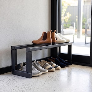 May include: A black wooden shoe rack with two horizontal tiers. The top tier holds a pair of tan leather Chelsea boots and a pair of white sneakers. The bottom tier holds a pair of grey knit sneakers, striped espadrilles, and dark blue loafers.