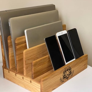 Può includere: Un organizer da scrivania in legno con più scomparti per dispositivi elettronici. Contiene diversi laptop, tablet e smartphone. L'organizer è realizzato in legno chiaro e presenta un emblema di leone sul davanti.