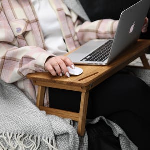 May include: A person using a laptop and a wireless mouse on a wooden lap desk. The desk is supported by two foldable legs. The person is sitting on a couch with a gray blanket.