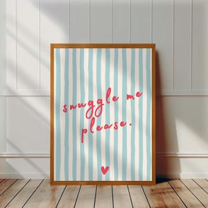 Op de afbeelding: Een ingelijste print met een lichtblauwe en witte gestreepte achtergrond. De print bevat de woorden "snuggle me please" in roze script, met een klein rood hartje onderaan. De lijst heeft een warme bruine kleur.