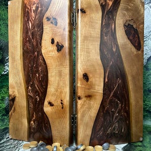 May include: Backgammon board made of wood and dark brown resin. The board is open, revealing the playing surface. A pile of game pieces in shades of gray and tan are in front of the board. The wood has a natural grain pattern.