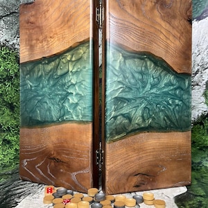May include: A wooden backgammon set with a teal and green epoxy resin river design. The set is open, revealing the playing surface, and includes game pieces and dice. The wood has a natural grain, and the set is hinged.