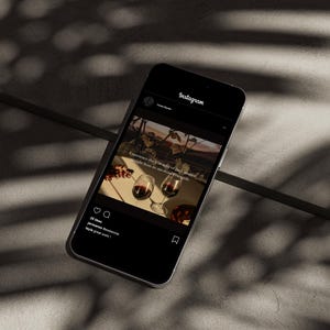 May include: A smartphone displays an Instagram post featuring two wine glasses filled with red wine. The post includes text about wine and a vineyard scene. The phone is on a textured surface with shadows.