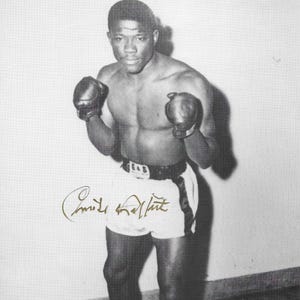 May include: Black and white photo of a boxer wearing boxing gloves and shorts. The boxer is standing in a boxing ring. The photo is signed "Emile Griffith".