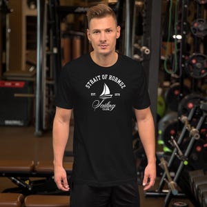 Op de afbeelding: Zwart T-shirt met een witte opdruk van "Strait of Hormuz Sailing Club" en een zeilboot illustratie. De tekst bevat "Est. 1979". Het shirt heeft korte mouwen en een ronde hals.