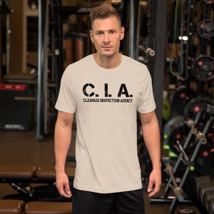 Op de afbeelding: Een lichtbeige t-shirt met de tekst "C.I.A. CLEAVAGE INSPECTION AGENCY" in het zwart. Het shirt heeft korte mouwen en een ronde hals. De persoon staat in een sportschool.